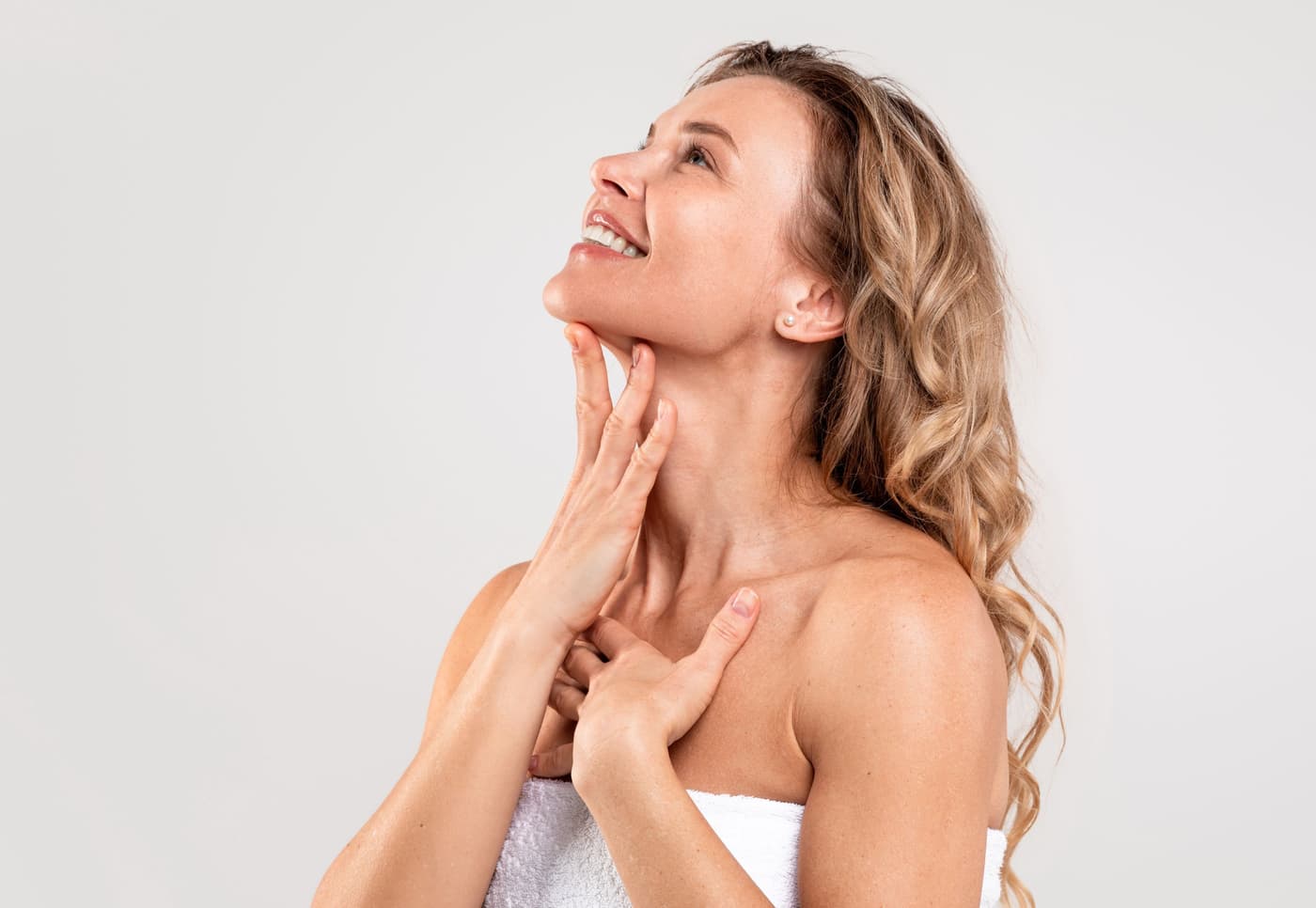 Lächelnde Frau mit sonnengebräunter Haut trägt sanft Feuchtigkeitscreme auf ihren Hals auf, helles Studiohintergrund, frische Sommerausstrahlung.